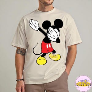 Puede incluir: Camiseta beige con un gr&aacute;fico de Mickey Mouse haciendo un dab. Mickey est&aacute; representado en negro, rojo, amarillo y blanco. La camiseta tiene un corte holgado y mangas cortas. El texto "ICMOSTORE" es visible en la parte inferior derecha.