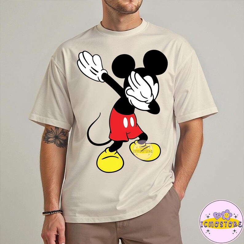 Puede incluir: Camiseta beige con un gr&aacute;fico de Mickey Mouse haciendo un dab. Mickey est&aacute; representado en negro, rojo, amarillo y blanco. La camiseta tiene un corte holgado y mangas cortas. El texto "ICMOSTORE" es visible en la parte inferior derecha.