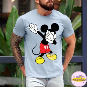 Puede incluir: Camiseta azul claro con un gr&aacute;fico de Mickey Mouse en pose de dab. Mickey est&aacute; representado en sus colores cl&aacute;sicos: negro, rojo, amarillo y blanco. La camiseta tiene mangas cortas y cuello redondo. El texto "ICMOSTORE" es visible en la parte inferior derecha.