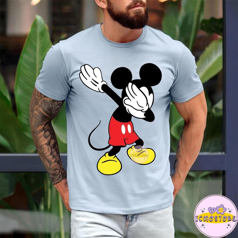Puede incluir: Camiseta azul claro con un gr&aacute;fico de Mickey Mouse en pose de dab. Mickey est&aacute; representado en sus colores cl&aacute;sicos: negro, rojo, amarillo y blanco. La camiseta tiene mangas cortas y cuello redondo. El texto "ICMOSTORE" es visible en la parte inferior derecha.