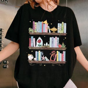 Puede incluir: Camiseta negra con una ilustraci&oacute;n caprichosa de tres estantes de libros coloridos y objetos de cuento de hadas. El dise&ntilde;o incluye una rana, una rosa en una campana de cristal, un drag&oacute;n y una l&aacute;mpara m&aacute;gica. Peque&ntilde;as estrellas est&aacute;n dispersas.
