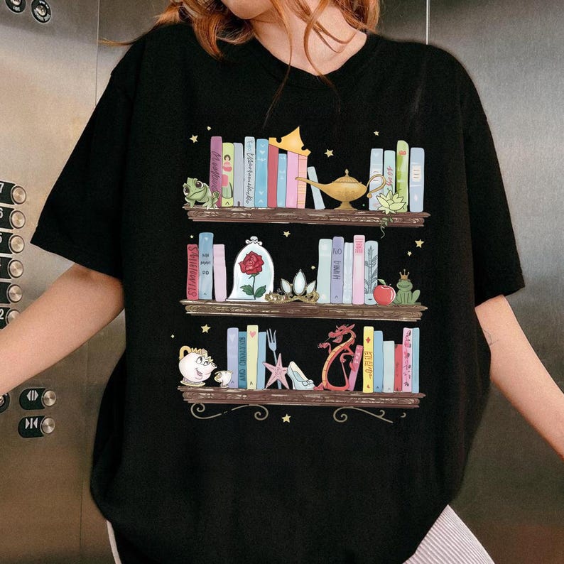 Puede incluir: Camiseta negra con una ilustraci&oacute;n caprichosa de tres estantes de libros coloridos y objetos de cuento de hadas. El dise&ntilde;o incluye una rana, una rosa en una campana de cristal, un drag&oacute;n y una l&aacute;mpara m&aacute;gica. Peque&ntilde;as estrellas est&aacute;n dispersas.