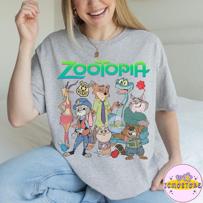 Pu&ograve; includere: Una maglietta grigia con la scritta "Zootopia" in verde e blu, con personaggi dei cartoni animati del film. Il design include un coniglio, una volpe e altri animali. La maglietta ha un girocollo.