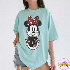May include: A light blue t-shirt featuring a graphic of Minnie Mouse. Minnie Mouse is depicted in a relaxed pose, wearing a red bow with white polka dots. The shirt is a comfortable, oversized fit.
