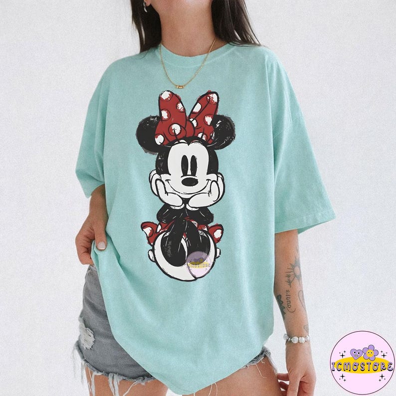May include: A light blue t-shirt featuring a graphic of Minnie Mouse. Minnie Mouse is depicted in a relaxed pose, wearing a red bow with white polka dots. The shirt is a comfortable, oversized fit.