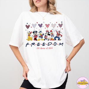 May include: White t-shirt featuring cartoon characters holding balloons in red, white, and blue. The word "FREEDOM" is printed in navy blue, with "250 Years of USA" below. The design includes stars and patriotic colors.