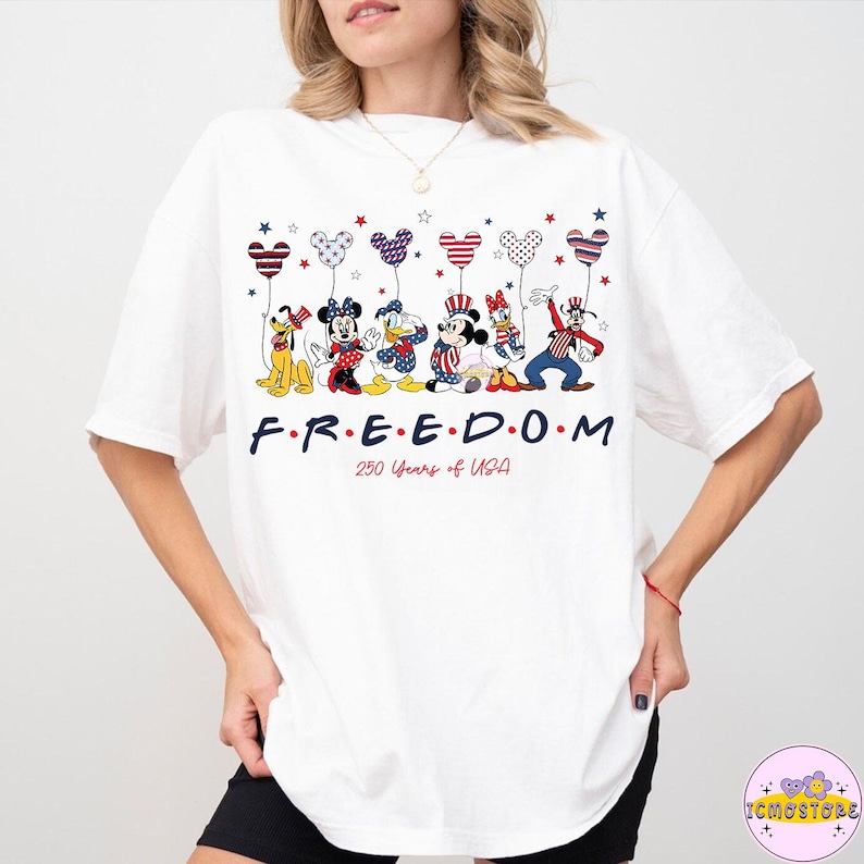 May include: White t-shirt featuring cartoon characters holding balloons in red, white, and blue. The word "FREEDOM" is printed in navy blue, with "250 Years of USA" below. The design includes stars and patriotic colors.