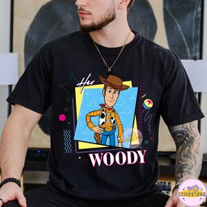May include: Black t-shirt featuring a graphic of Woody from Toy Story. The design includes the text "Her" above the character and "WOODY" below. The graphic is set against a blue sky background with a yellow and pink frame.