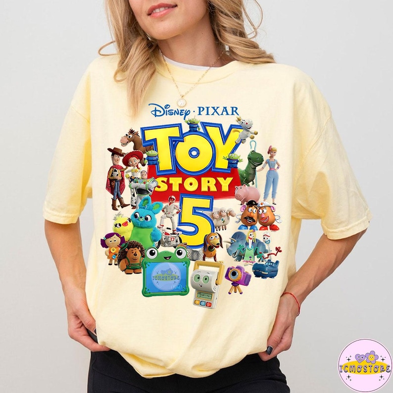May include: A light yellow t-shirt featuring the Disney Pixar Toy Story 5 logo with various character illustrations. The design includes the words "TOY STORY 5" in bold, colorful letters, surrounded by images of Woody, Buzz Lightyear, and other characters.