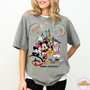 May include: Gray t-shirt featuring a colorful Disney graphic with Mickey Mouse, Minnie Mouse, Donald Duck, Goofy, and other characters. The shirt includes the text "Disney Family Vacation" and the year "2026".