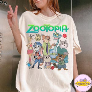 Pu&ograve; includere: T-shirt color crema con la scritta "ZOOTOPIA" in verde e blu, con una grafica di vari personaggi dei cartoni animati. I personaggi includono un coniglio in uniforme da poliziotto, una volpe e un serpente.