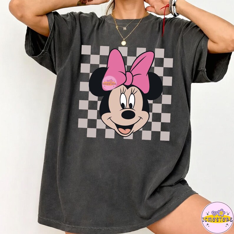 Puede incluir: Camiseta gris oscuro con un dise&ntilde;o de Minnie Mouse con un lazo rosa y un fondo a cuadros. El dise&ntilde;o est&aacute; centrado en la camiseta. La camiseta tiene mangas cortas y un corte holgado. El texto "ICMOSTORE" est&aacute; en la parte inferior derecha.