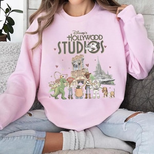 Puede incluir: Sudadera rosa claro con el texto "Disney's Hollywood Studios" y un gráfico de personajes de Toy Story y atracciones del parque. El diseño incluye un dinosaurio verde, un vaquero y un guardabosques espacial.