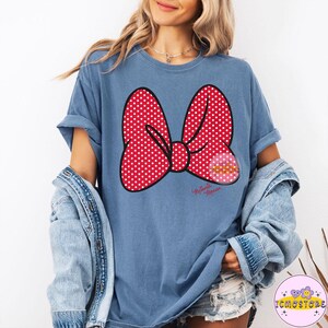 May include: Blue t-shirt featuring a large red bow with white polka dots. The bow has a black outline and the words "Minnie Mouse" are printed in cursive below the bow. A denim jacket is draped over the shoulders.