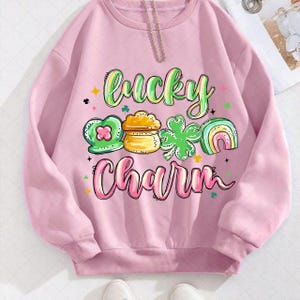 Puede incluir: Una sudadera rosa claro con las palabras "Lucky Charm" en una fuente colorida y caprichosa. El diseño incluye un trébol verde, una olla de oro y un arcoíris, todo en un estilo lúdico, perfecto para el Día de San Patricio.