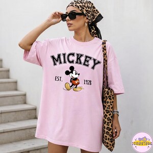 May include: A light pink t-shirt featuring the word "MICKEY" in black block letters above a graphic of Mickey Mouse. Below the graphic are the words "EST. 1928". The person is wearing sunglasses and a leopard print scarf and carrying a leopard print tote bag.