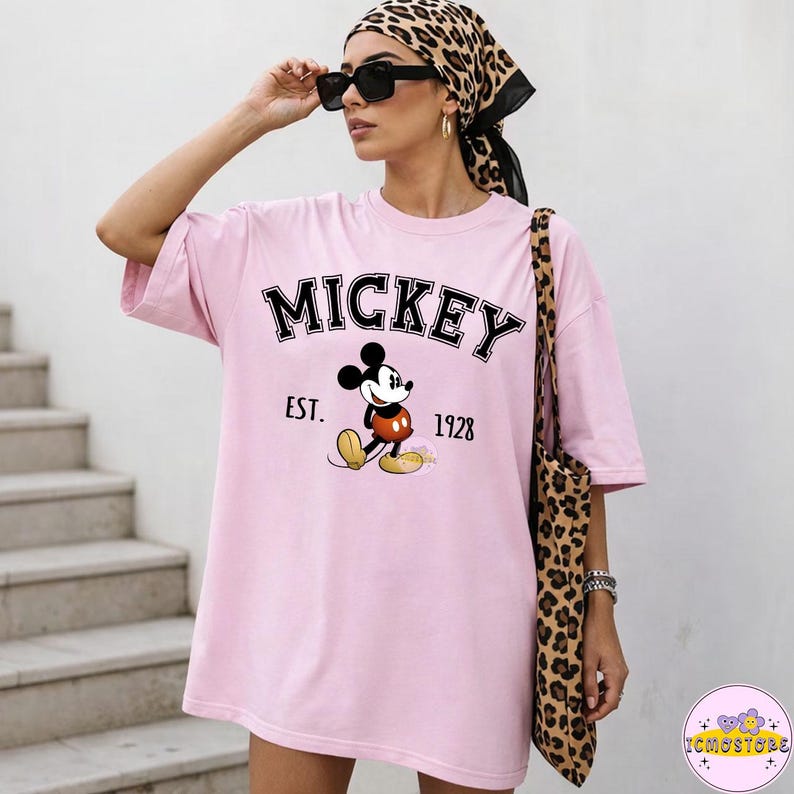 May include: A light pink t-shirt featuring the word "MICKEY" in black block letters above a graphic of Mickey Mouse. Below the graphic are the words "EST. 1928". The person is wearing sunglasses and a leopard print scarf and carrying a leopard print tote bag.