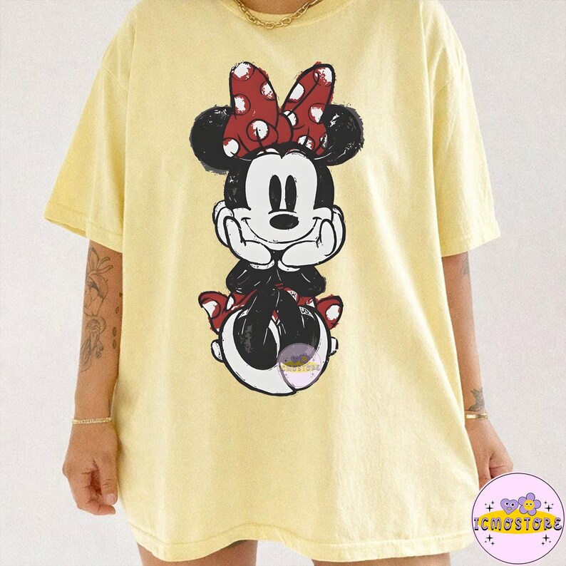May include: Yellow t-shirt featuring a distressed graphic of Minnie Mouse. The cartoon character is depicted with a red and white polka dot bow, black ears, and a smiling face. The shirt has short sleeves and a relaxed fit.