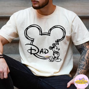 May include: Cream-colored t-shirt featuring a black outline of Mickey Mouse ears with the word "DAD" in a stylized font. A small sketch of Mickey Mouse is seated below the text. The shirt is a casual style.