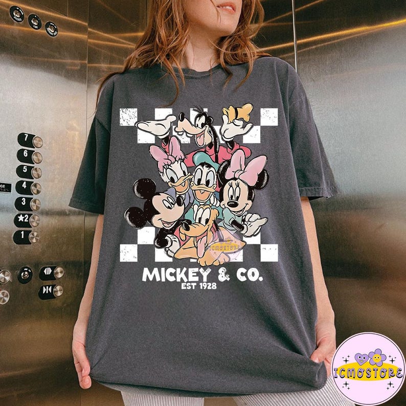 May include: Dark gray t-shirt featuring a graphic of Mickey Mouse and friends, including Minnie Mouse, Donald Duck, and Goofy, on a white and black checkered background. The text "MICKEY & CO. EST 1928" is printed below the characters. The shirt is a casual wear item.