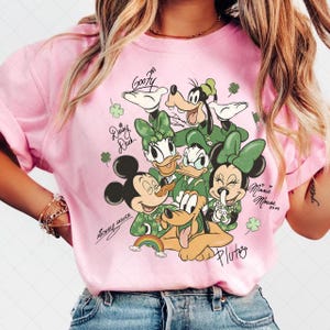Puede incluir: Camiseta rosa con un gráfico de los personajes de Disney Mickey Mouse, Minnie Mouse, Donald Duck, Daisy Duck, Goofy y Pluto, todos con trajes verdes. La camiseta incluye los nombres de los personajes y un arcoíris.