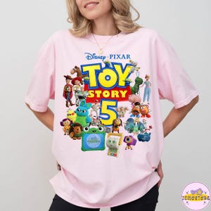May include: A light pink t-shirt featuring a colorful graphic of characters from the Disney Pixar movie Toy Story 5. The design includes the movie title and the number 5 in yellow and red.