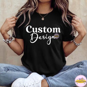 May include: Black t-shirt with the words "Custom Design" in white script. The shirt has a crew neck and short sleeves. The image also includes a logo in the lower right corner.