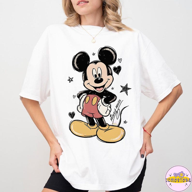 May include: White t-shirt featuring a cartoon illustration of Mickey Mouse. The design includes Mickey in his classic outfit with red shorts, yellow shoes, and black ears. Surrounding Mickey are black hearts and stars.