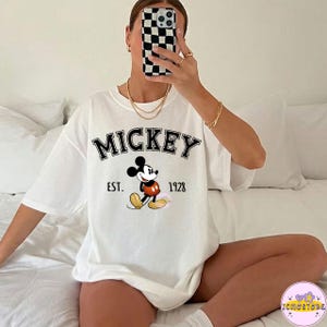 May include: White t-shirt featuring the word "MICKEY" in black, with "EST. 1928" below. A cartoon Mickey Mouse is in the center, wearing red shorts and yellow shoes. The shirt is a casual, oversized style.
