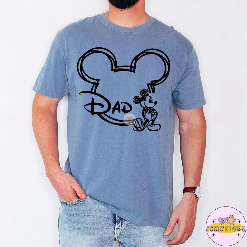 May include: A blue t-shirt featuring a black graphic of Mickey Mouse ears with the word "Dad" and a small Mickey Mouse figure. The shirt is a solid blue color.