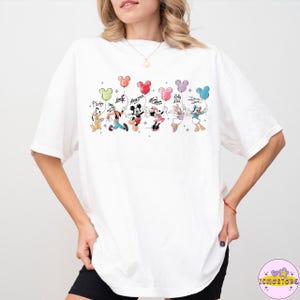 以下が含まれることがあります： ミッキーマウスの形をした風船を持っているディズニーキャラクターのカラフルなグラフィックが特徴の白いTシャツ。キャラクターには、プルート、グーフィー、ミッキー、ミニー、デイジー、ドナルドダックが含まれます。キャラクターの名前が上に書かれています。