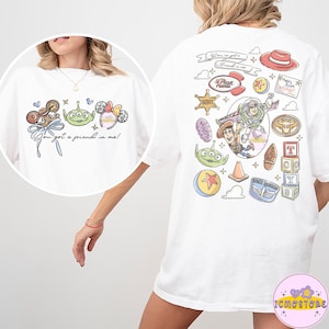 以下が含まれることがあります： カラフルな漫画のグラフィックとテキストが特徴の白いTシャツ。前面にはキャラクターと「You got a friend in me！」というフレーズが表示されています。背面には、「Andy」や「Room Keep Out」など、追加のキャラクターとフレーズが表示されています。