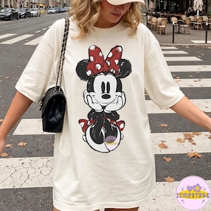 May include: Cream-colored oversized t-shirt featuring a distressed graphic of Minnie Mouse. Minnie is depicted with a red bow with white polka dots. The graphic is in black, red, and white. The shirt has short sleeves.