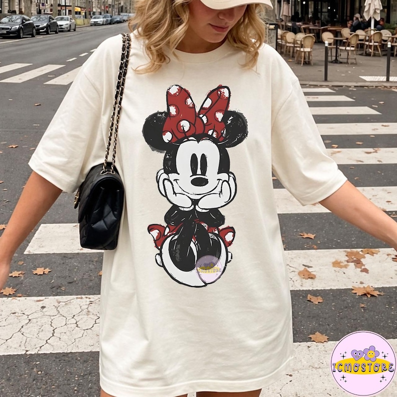May include: Cream-colored oversized t-shirt featuring a distressed graphic of Minnie Mouse. Minnie is depicted with a red bow with white polka dots. The graphic is in black, red, and white. The shirt has short sleeves.