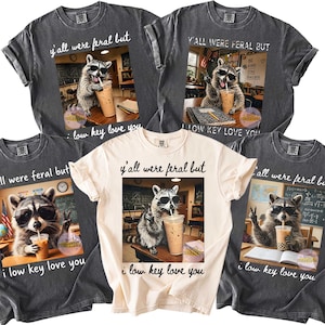 Puede incluir: Cinco camisetas con un diseño de mapache con gafas de sol bebiendo de un vaso. Las camisetas son de varios colores, incluyendo gris y crema, con el texto "y'all were feral but i low key love you".