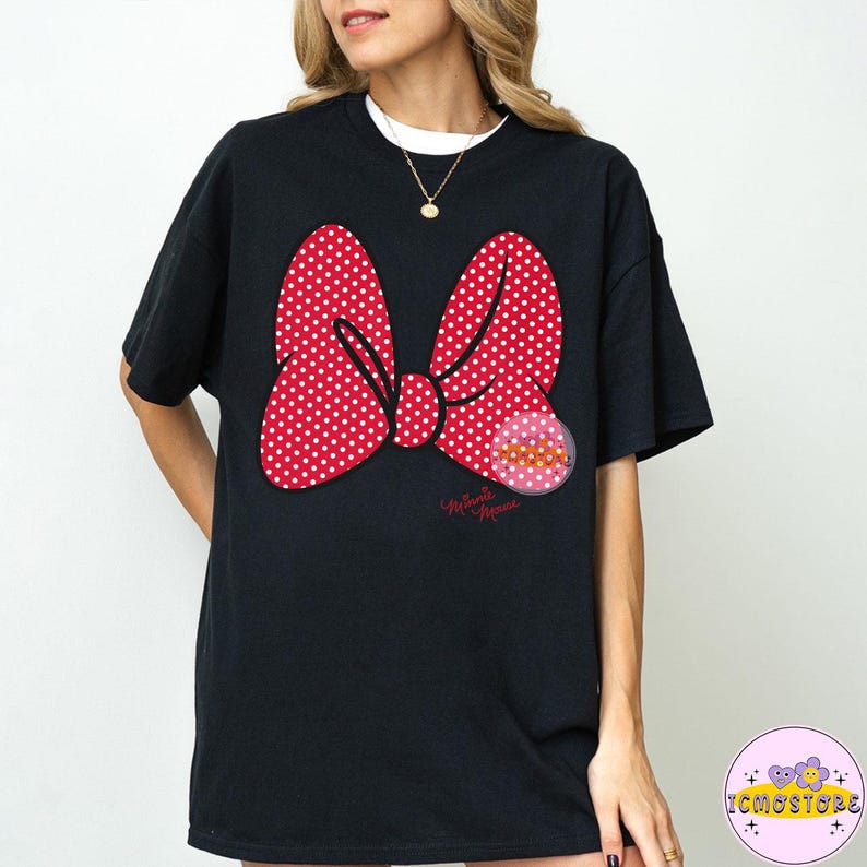 May include: Black t-shirt featuring a large red bow with white polka dots. A pink circular graphic with text is on the right side of the bow. The words "Minnie Mouse" are written in cursive below the bow.