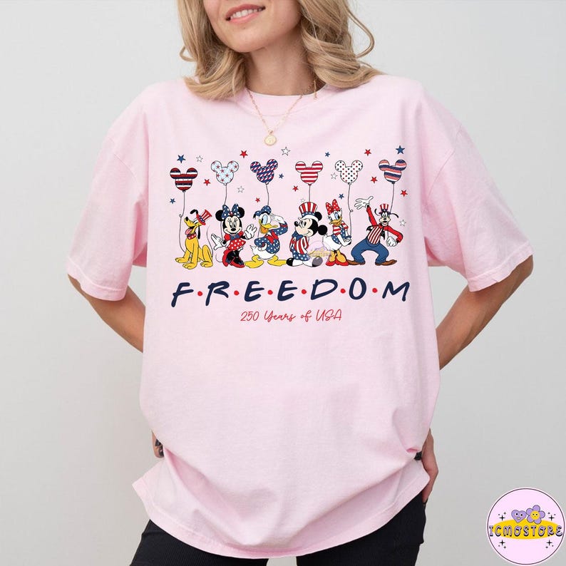 May include: A light pink t-shirt featuring a graphic of Disney characters holding balloon-shaped fireworks. The word "FREEDOM" is printed in navy blue, with "250 Years of USA" below. The shirt is a relaxed fit.