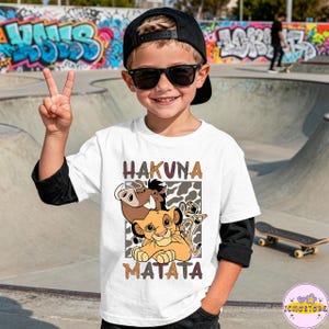 May include: White t-shirt with a graphic of Simba, Pumbaa, and Timon from The Lion King, with the words "HAKUNA MATATA" in brown letters. The shirt has black sleeves and is worn by a child wearing sunglasses and a black cap.