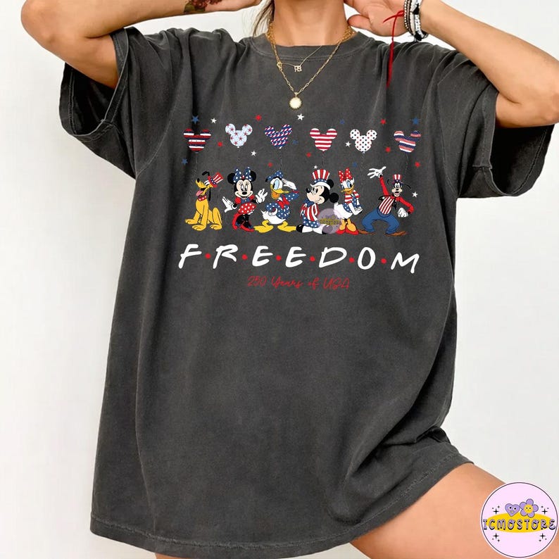 May include: Dark gray t-shirt featuring a graphic of Disney characters in patriotic attire. The word "FREEDOM" is printed in white, with "246 Years of U.S." below. Mickey, Minnie, Donald, Daisy, and Goofy are depicted in red, white, and blue.