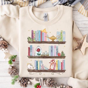 Puede incluir: Sudadera beige con una ilustraci&oacute;n caprichosa de tres estanter&iacute;as llenas de libros coloridos y objetos tem&aacute;ticos de cuentos de hadas. El dise&ntilde;o incluye una rana, una rosa en una c&uacute;pula de cristal, una l&aacute;mpara m&aacute;gica y un drag&oacute;n.