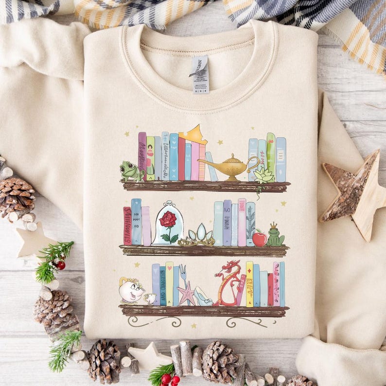 Puede incluir: Sudadera beige con una ilustraci&oacute;n caprichosa de tres estanter&iacute;as llenas de libros coloridos y objetos tem&aacute;ticos de cuentos de hadas. El dise&ntilde;o incluye una rana, una rosa en una c&uacute;pula de cristal, una l&aacute;mpara m&aacute;gica y un drag&oacute;n.