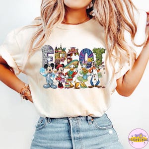 May include: Cream-colored t-shirt featuring the word "EPCOT" in colorful, themed lettering. The letters are filled with images of Disney characters and landmarks. Mickey Mouse, Minnie Mouse, Donald Duck, Daisy Duck, and Goofy are also depicted.