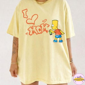 Könnte beinhalten: Hellgelbes T-Shirt mit rotem Graffiti-Schriftzug "I ❤️ Mom". Eine Zeichentrickfigur hält eine Sprühdose. Das Shirt hat eine lockere, übergroße Passform für den Alltag.