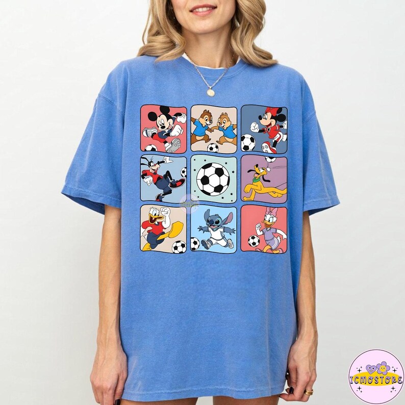 May include: A light blue t-shirt featuring a grid of cartoon characters playing soccer. The characters include Mickey Mouse, Chip and Dale, and Donald Duck. The shirt has a crew neck and short sleeves.