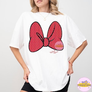 May include: White t-shirt featuring a large red polka-dot bow graphic with a smaller pink and yellow design. The bow has a black outline and the words "Minnie Mouse" are written in cursive below the bow.