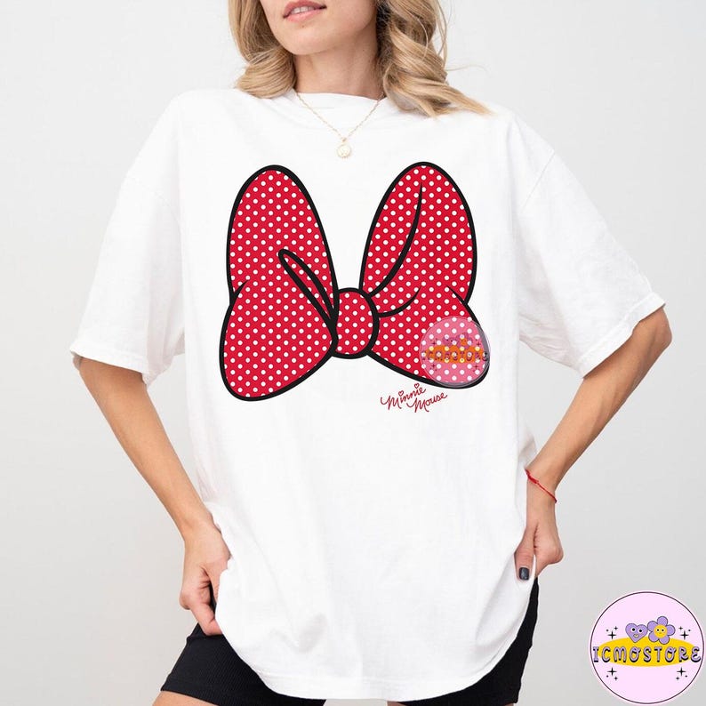 May include: White t-shirt featuring a large red polka-dot bow graphic with a smaller pink and yellow design. The bow has a black outline and the words "Minnie Mouse" are written in cursive below the bow.