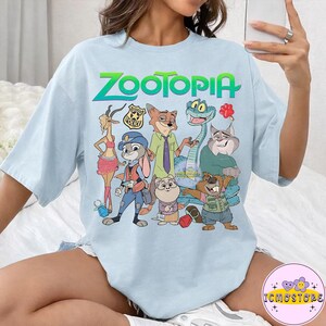 Pu&ograve; includere: T-shirt azzurra con la scritta "ZOOTOPIA" in verde e blu, e una grafica dei personaggi del film d'animazione. I personaggi includono una volpe, un coniglio, un serpente e altri animali. La maglietta ha un girocollo e maniche corte.