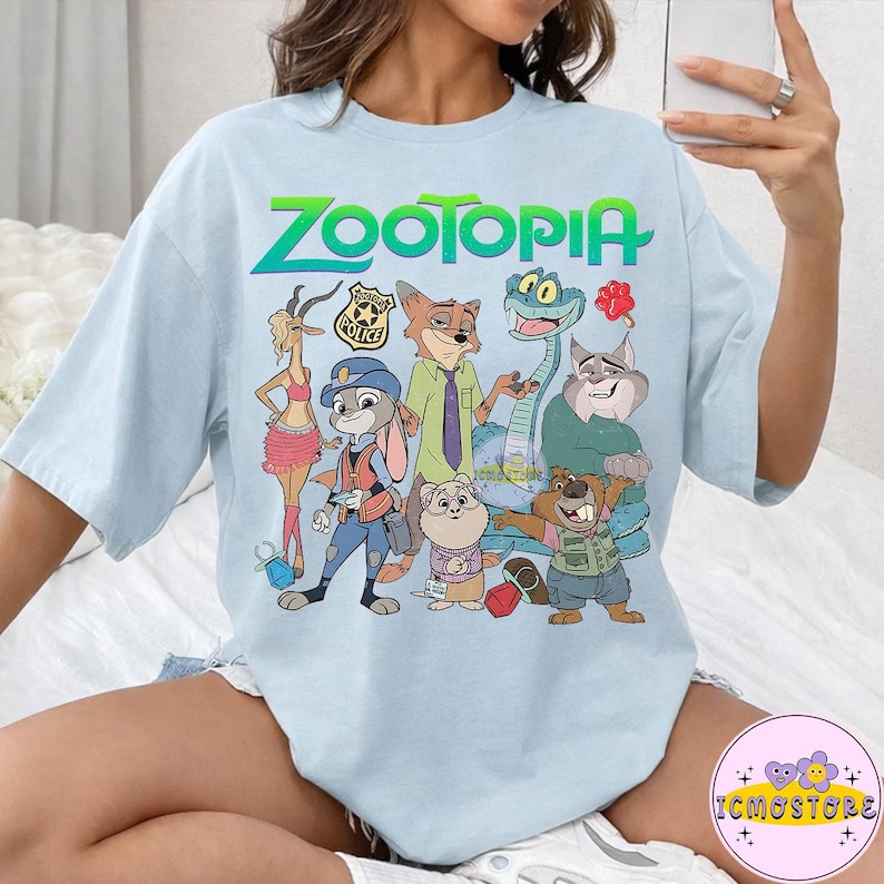 Pu&ograve; includere: T-shirt azzurra con la scritta "ZOOTOPIA" in verde e blu, e una grafica dei personaggi del film d'animazione. I personaggi includono una volpe, un coniglio, un serpente e altri animali. La maglietta ha un girocollo e maniche corte.