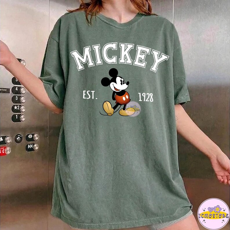 May include: Green t-shirt featuring the word "MICKEY" in white block letters above a cartoon image of Mickey Mouse. The text "EST. 1928" is printed below the character. The shirt has short sleeves and a relaxed fit.