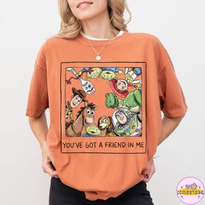 以下が含まれることがあります： オレンジ色のTシャツ。映画「トイ・ストーリー」のキャラクターのカラフルなイラストが描かれています。 ウッディ、バズ・ライトイヤー、その他のキャラクターが描かれており、下には「YOU'VE GOT A FRIEND IN ME」の文字があります。